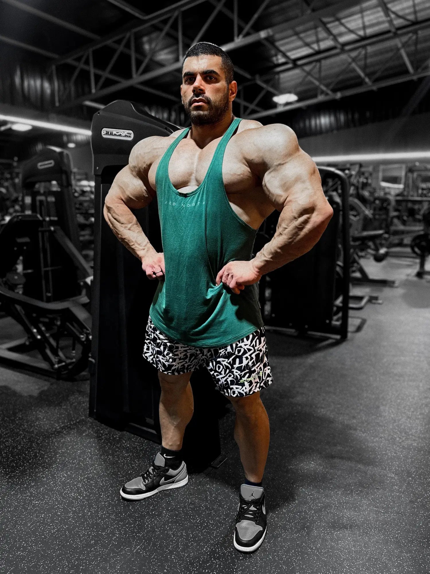 A muscular man wearing a sea green classic bodybuilding racerback stringer and black-and-white shorts in a gym