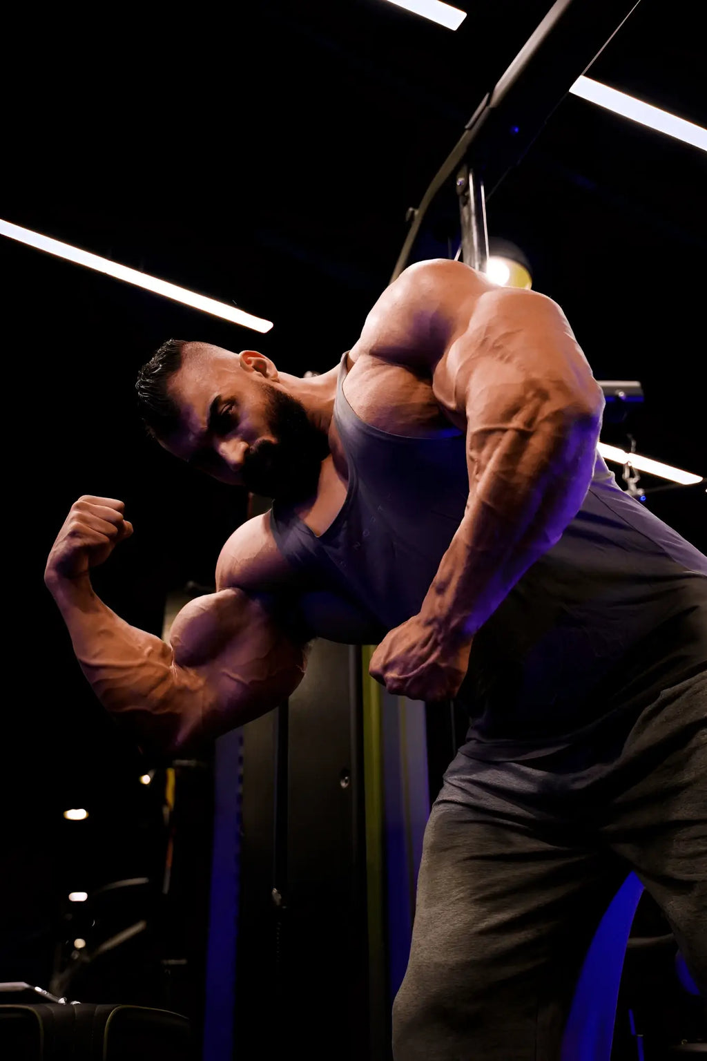 A muscular man flexes biceps wearing Jed North classic bodybuilding racerback stringer steel gray under gym lighting