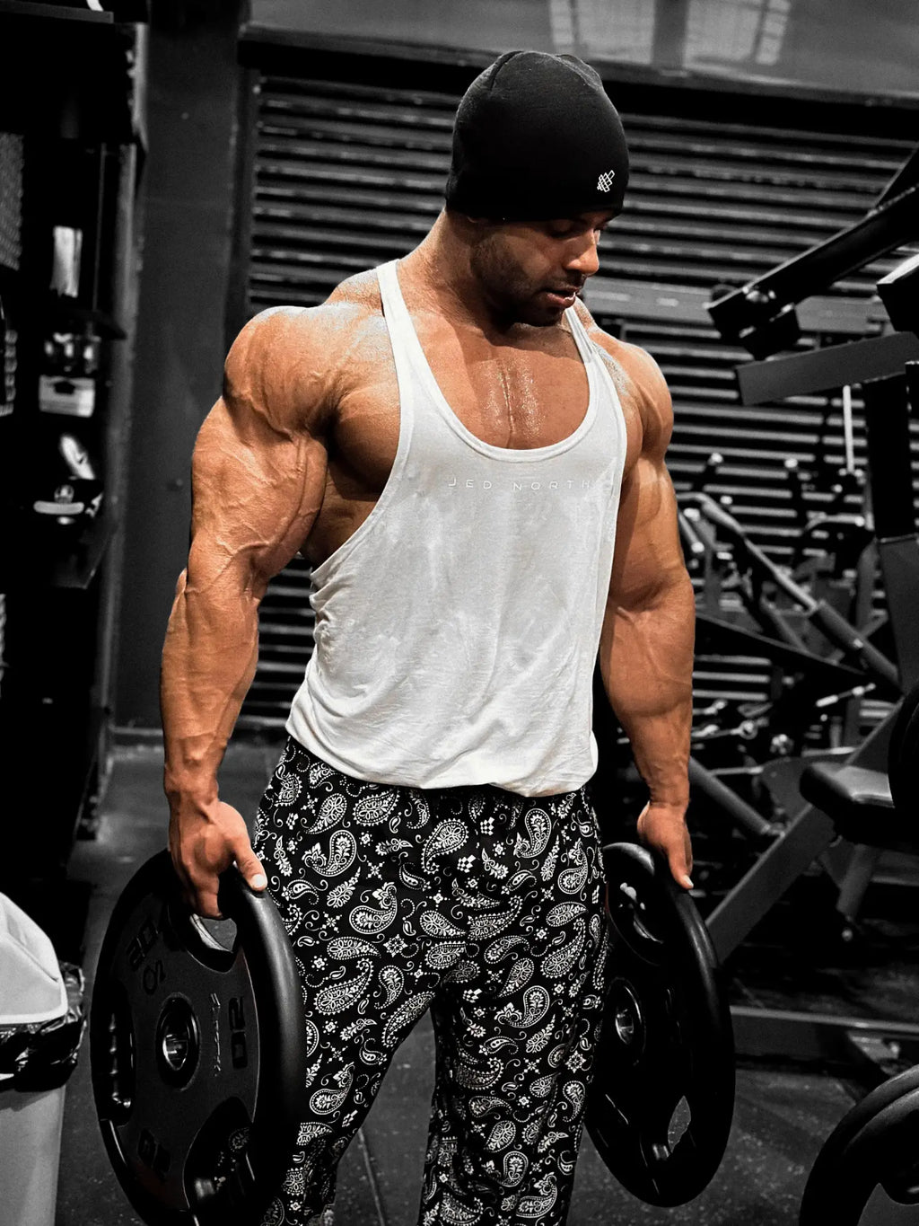 A muscular man wearing a white classic bodybuilding racerback stringer tank top and black paisley pants holding weight plates
