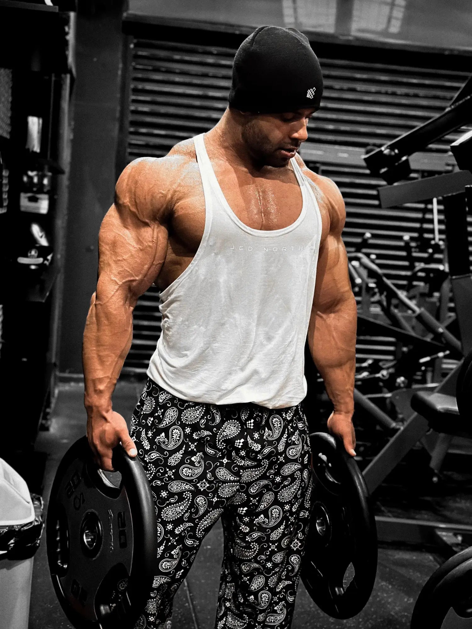 A muscular man wearing a white classic bodybuilding racerback stringer tank top and black paisley pants holding weight plates
