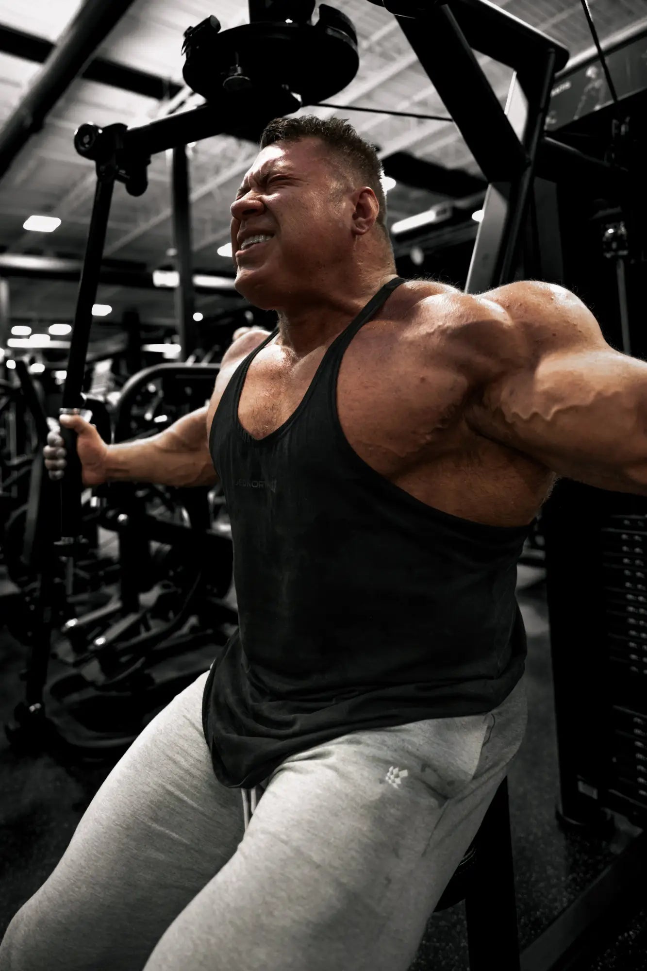 A muscular man wearing a black Jed North classic vintage washed racerback stringer in charcoal mid-exercise at the gym