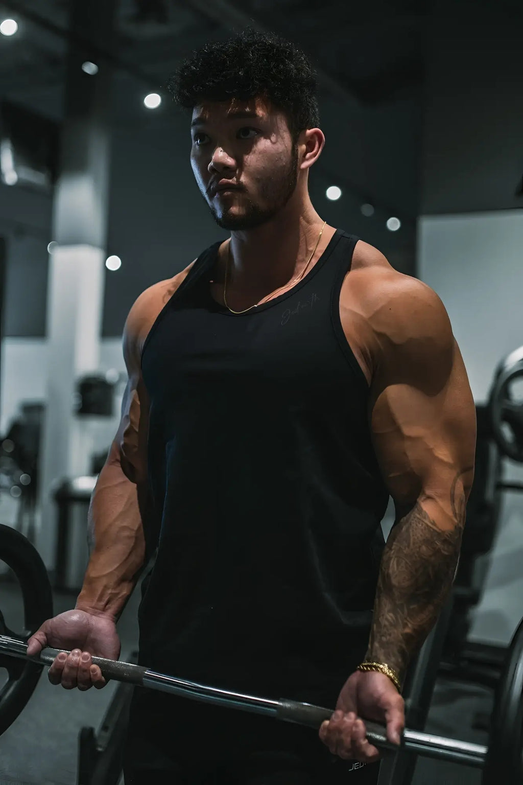 A muscular man wearing a black cotton training stringer tank top holding a barbell in a gym setting