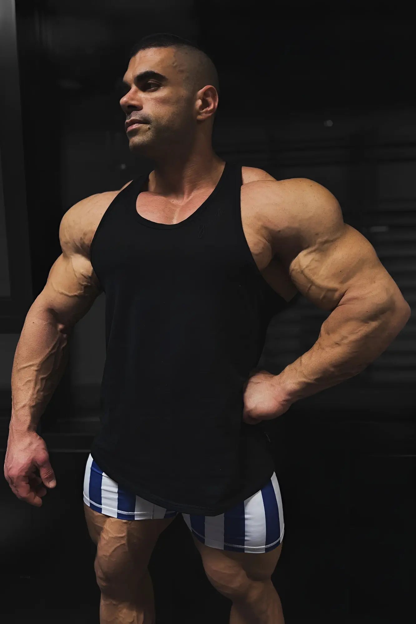 A muscular man wearing a black cotton training stringer tank top in midnight color and blue and white striped shorts