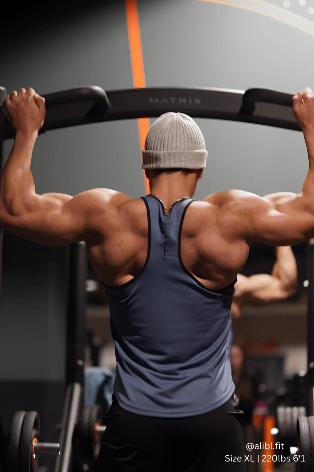 A man wearing a gray Jed North fast-dry bodybuilding workout stringer with black trim exercises on a gym machine