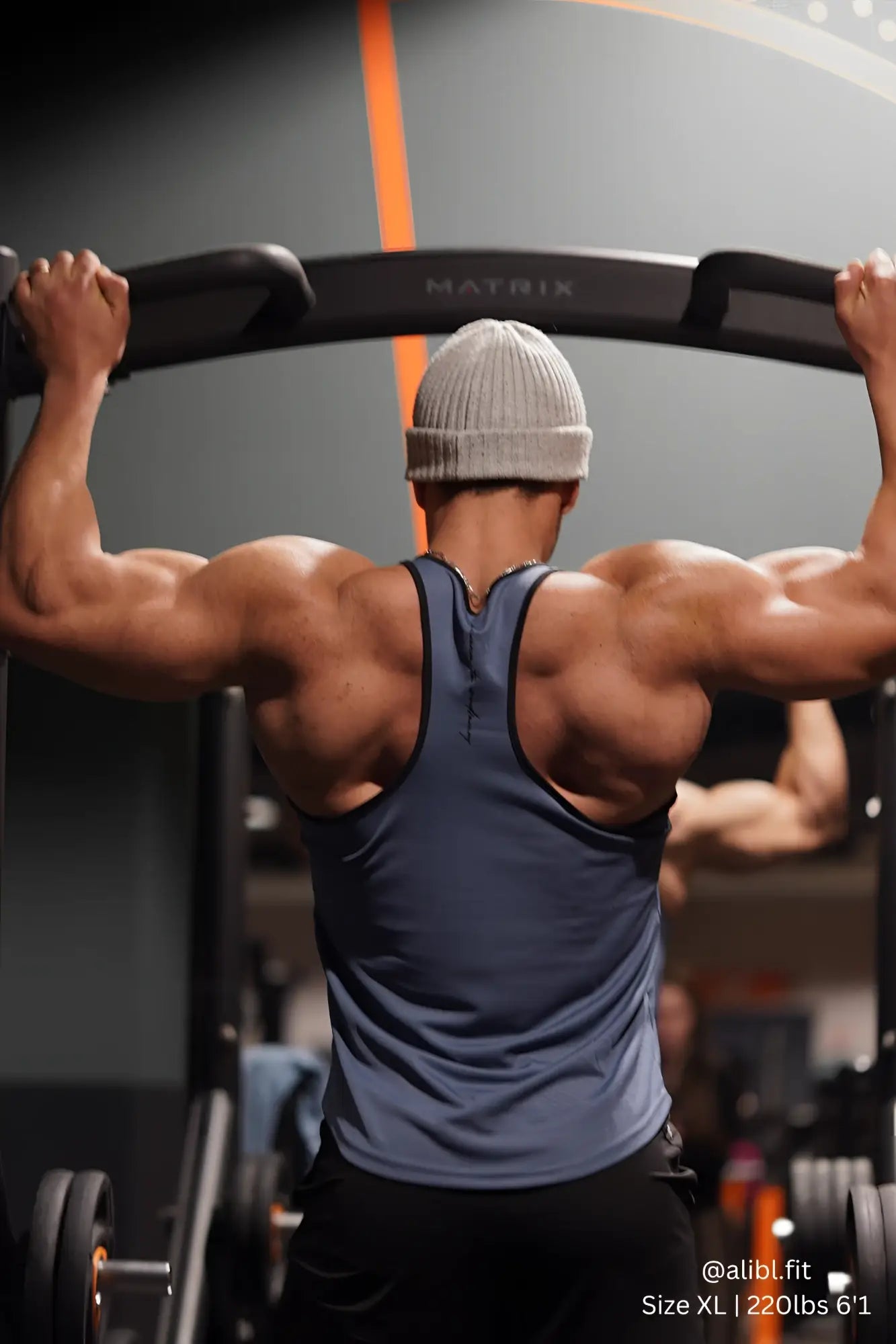 A man wearing a gray Jed North fast-dry bodybuilding workout stringer with black trim exercises on a gym machine