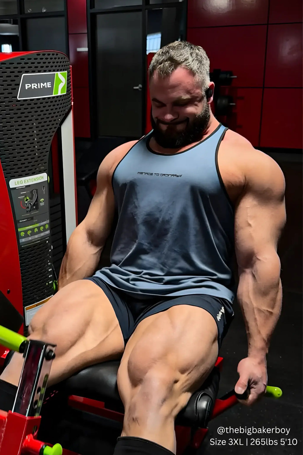 A muscular man wearing a gray and black Jed North fast-dry bodybuilding workout stringer seated on a red gym machine