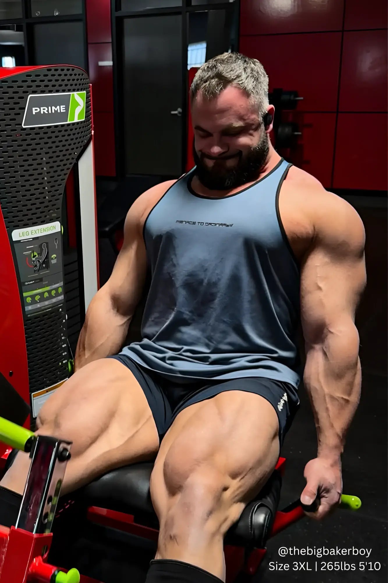 A muscular man wearing a gray and black Jed North fast-dry bodybuilding workout stringer seated on a red gym machine