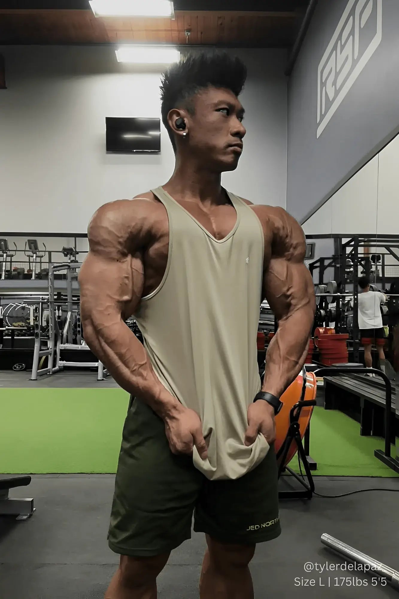 A muscular man wearing a khaki jed north fast-dry bodybuilding workout stringer tank top and dark green shorts