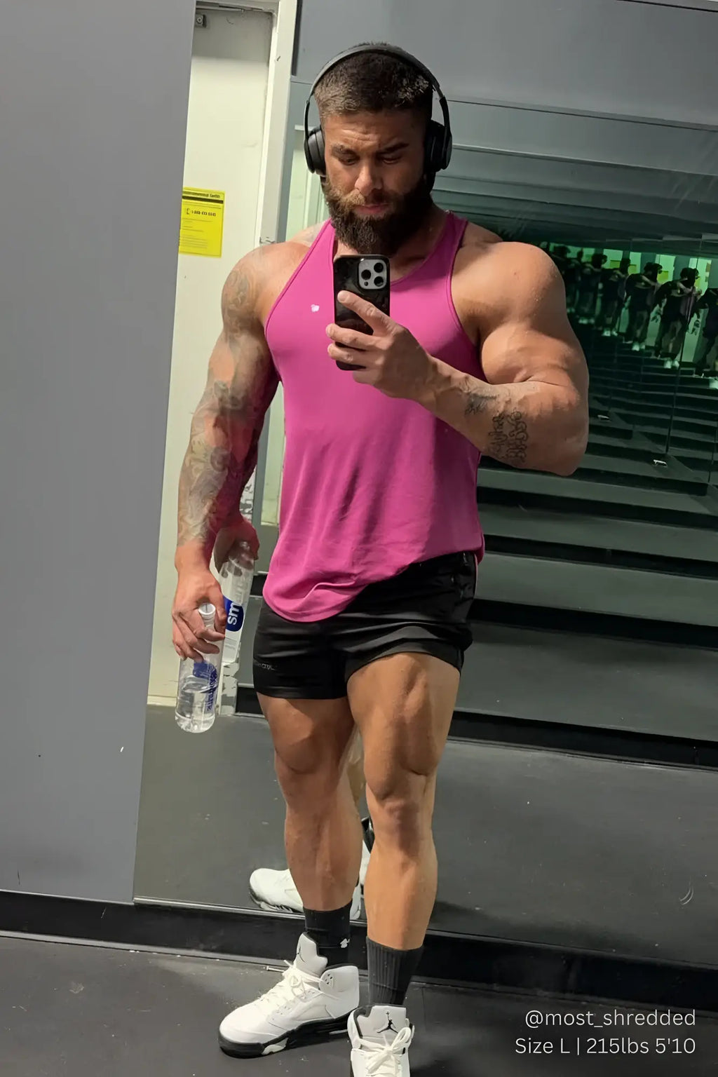 A muscular man wearing a fast-dry magenta sleeveless bodybuilding workout stringer holding a water bottle and taking a selfie