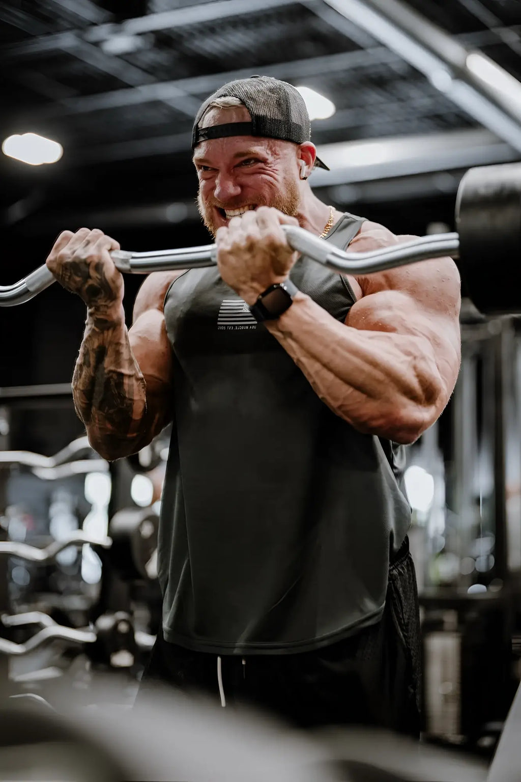 A muscular man wearing a black fast-dry bodybuilding workout stringer and cap grips a silver barbell in a gym