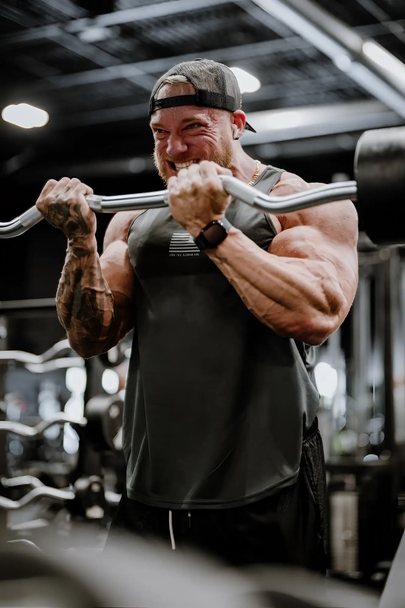 A muscular man wearing a black fast-dry bodybuilding workout stringer and cap grips a silver barbell in a gym