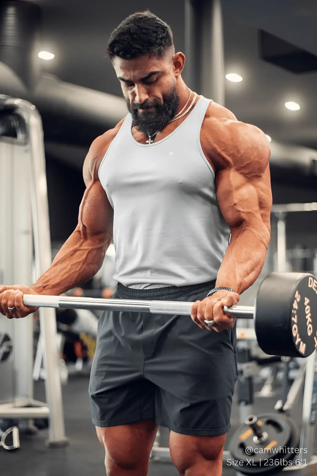 A man wearing a silver jed north fast-dry bodybuilding workout stringer holds a barbell in a gym setting