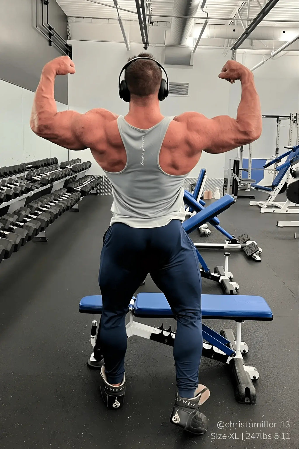 A muscular man in a silver jed north fast-dry bodybuilding workout stringer flexes arms in a gym