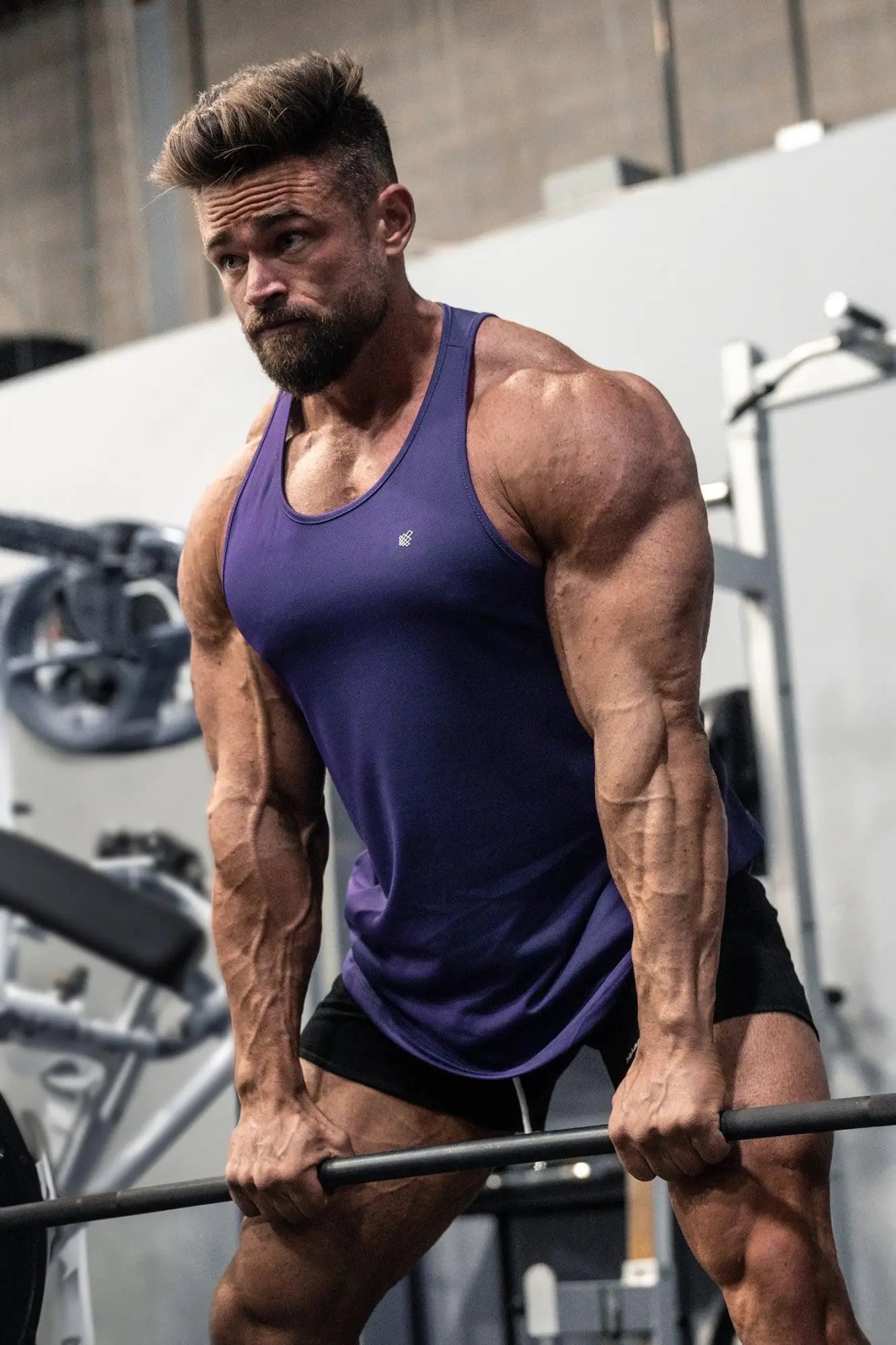 A muscular man wearing a violet jed north fast-dry bodybuilding workout stringer and black shorts gripping a barbell