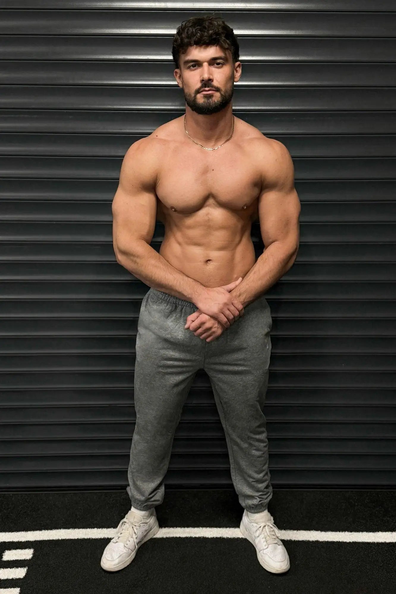 A muscular man wearing gray Jed North French Terry athletic training joggers and white sneakers stands confidently