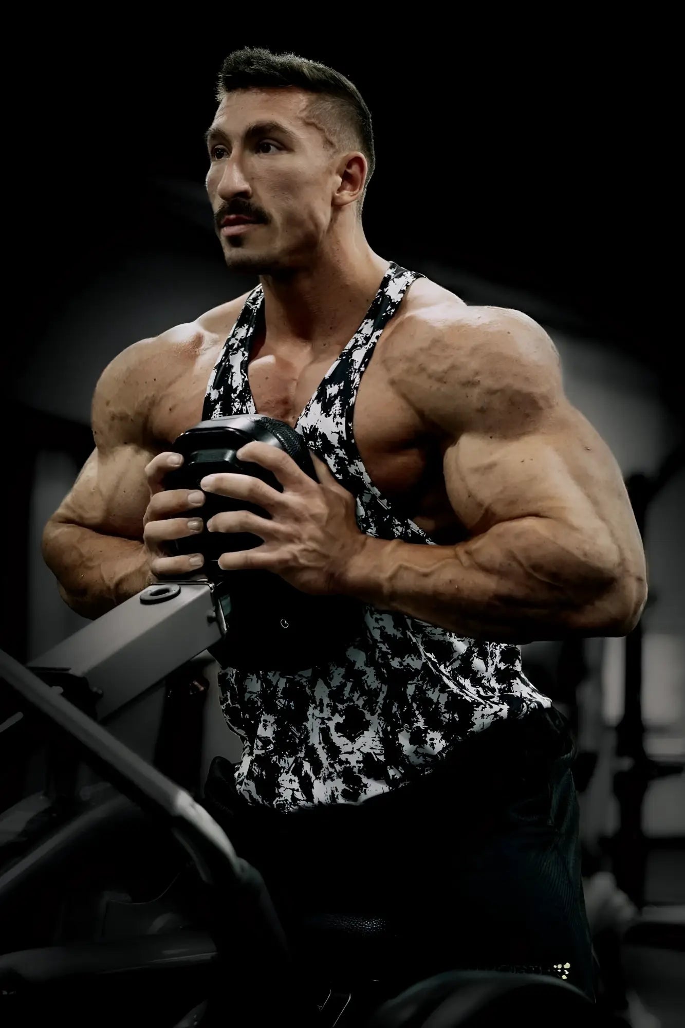 A muscular man wearing a black and white patterned Jed North graphic muscle stringer tank top holding a dumbbell