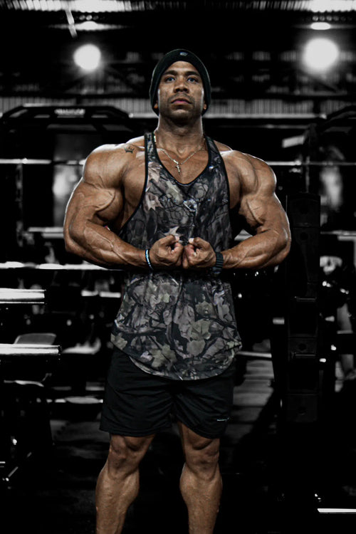Muscular man wearing jed north graphic muscle stringer tank top in hunter camo with black shorts front view