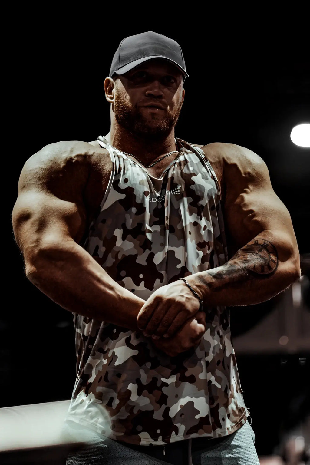 A man wearing a jed north graphic muscle stringer tank top in khaki camo with black and white splotches and a dark baseball cap