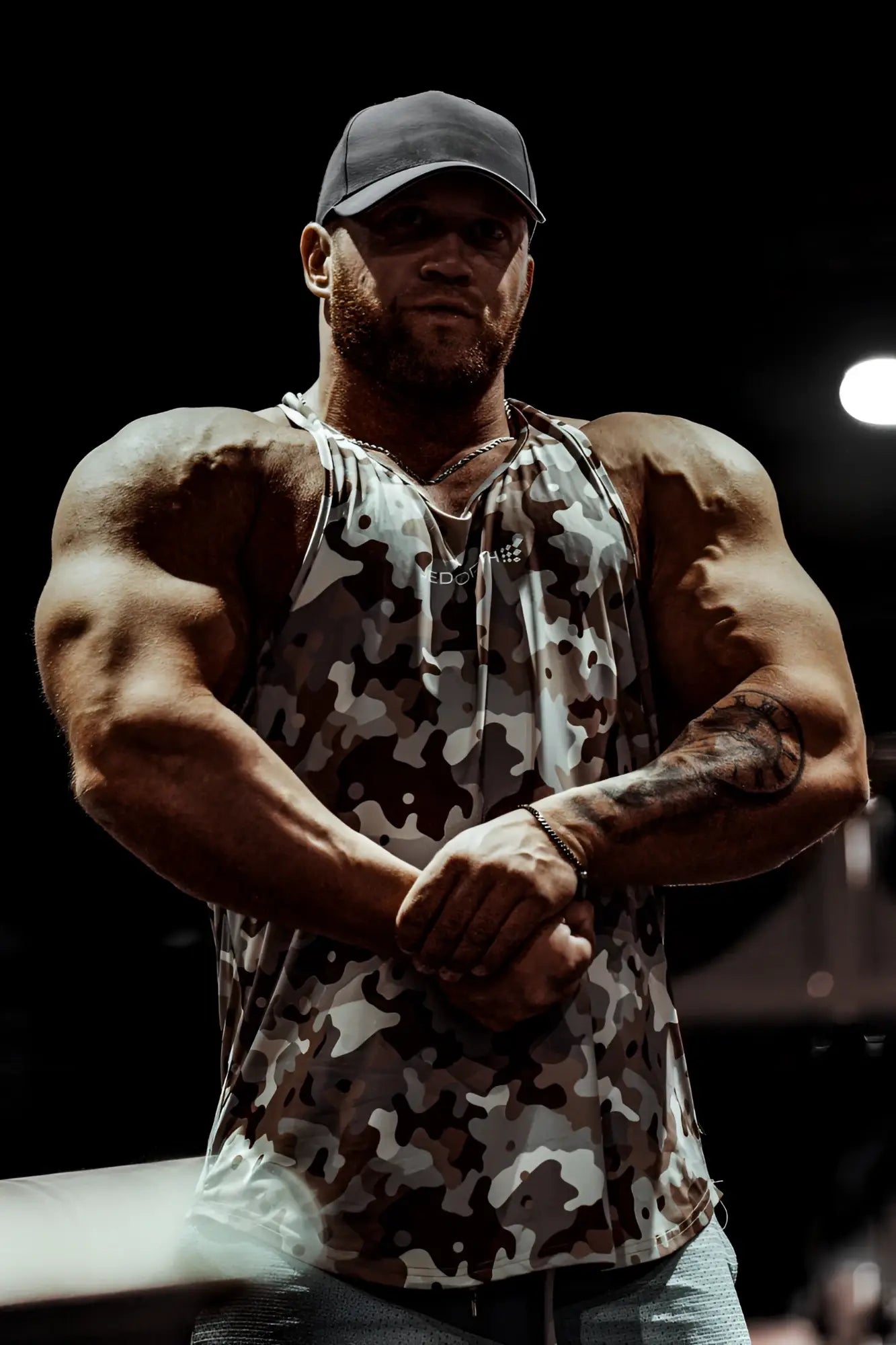 A man wearing a jed north graphic muscle stringer tank top in khaki camo with black and white splotches and a dark baseball cap