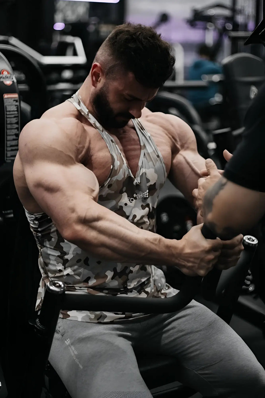 A muscular man wearing a khaki camo jed north graphic muscle stringer tank top seated on gym equipment