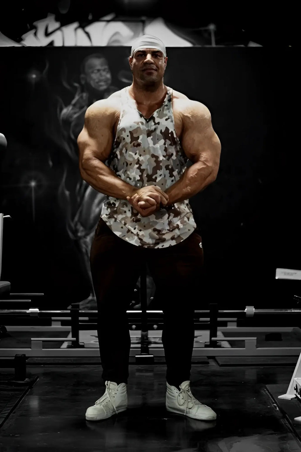 A muscular man wears a khaki camo Jed North graphic muscle stringer tank top in a gym setting
