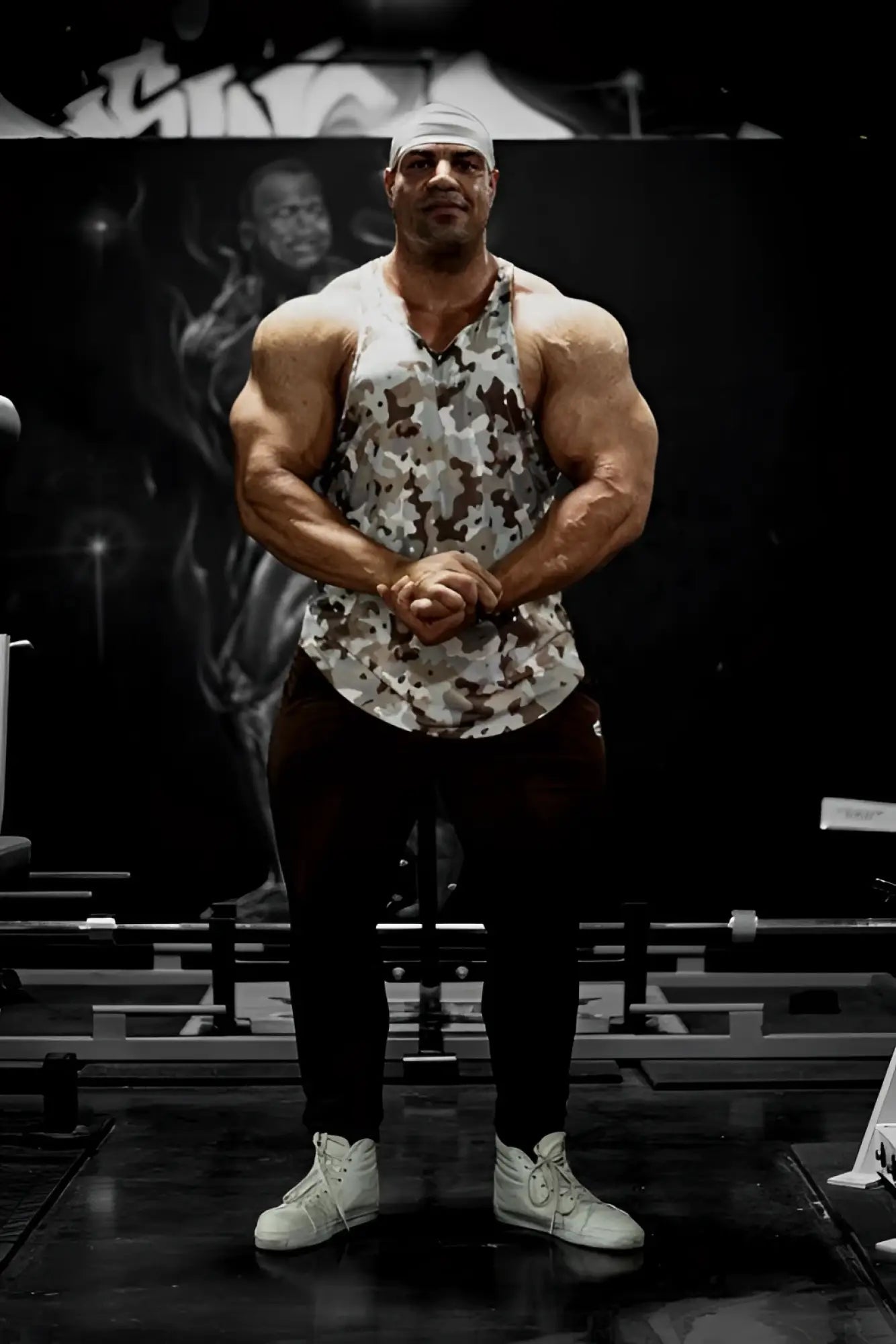 A muscular man wears a khaki camo Jed North graphic muscle stringer tank top in a gym setting