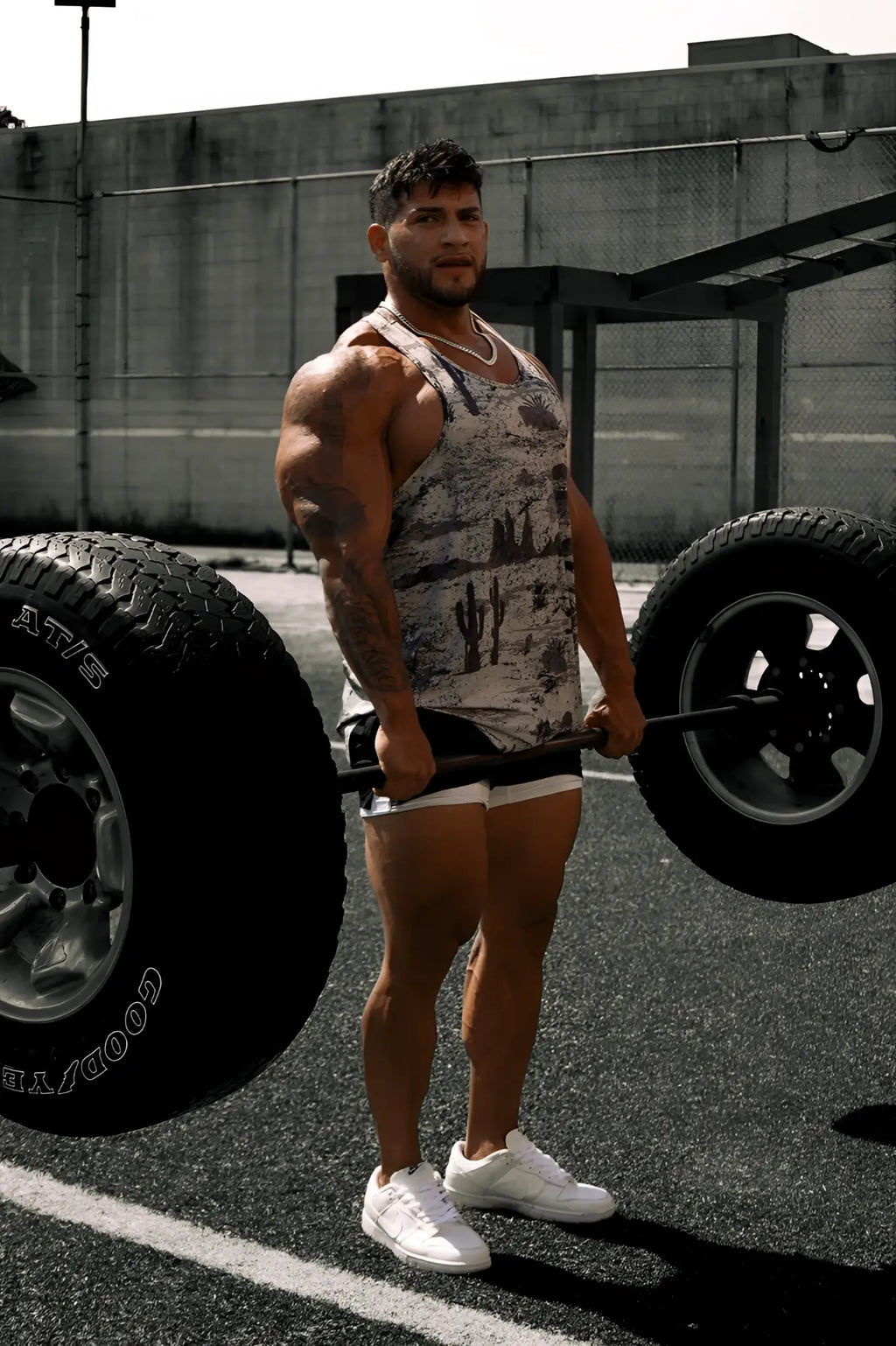 A muscular man wearing a gray and white cactus patterned graphic muscle stringer lifting a barbell with large tires