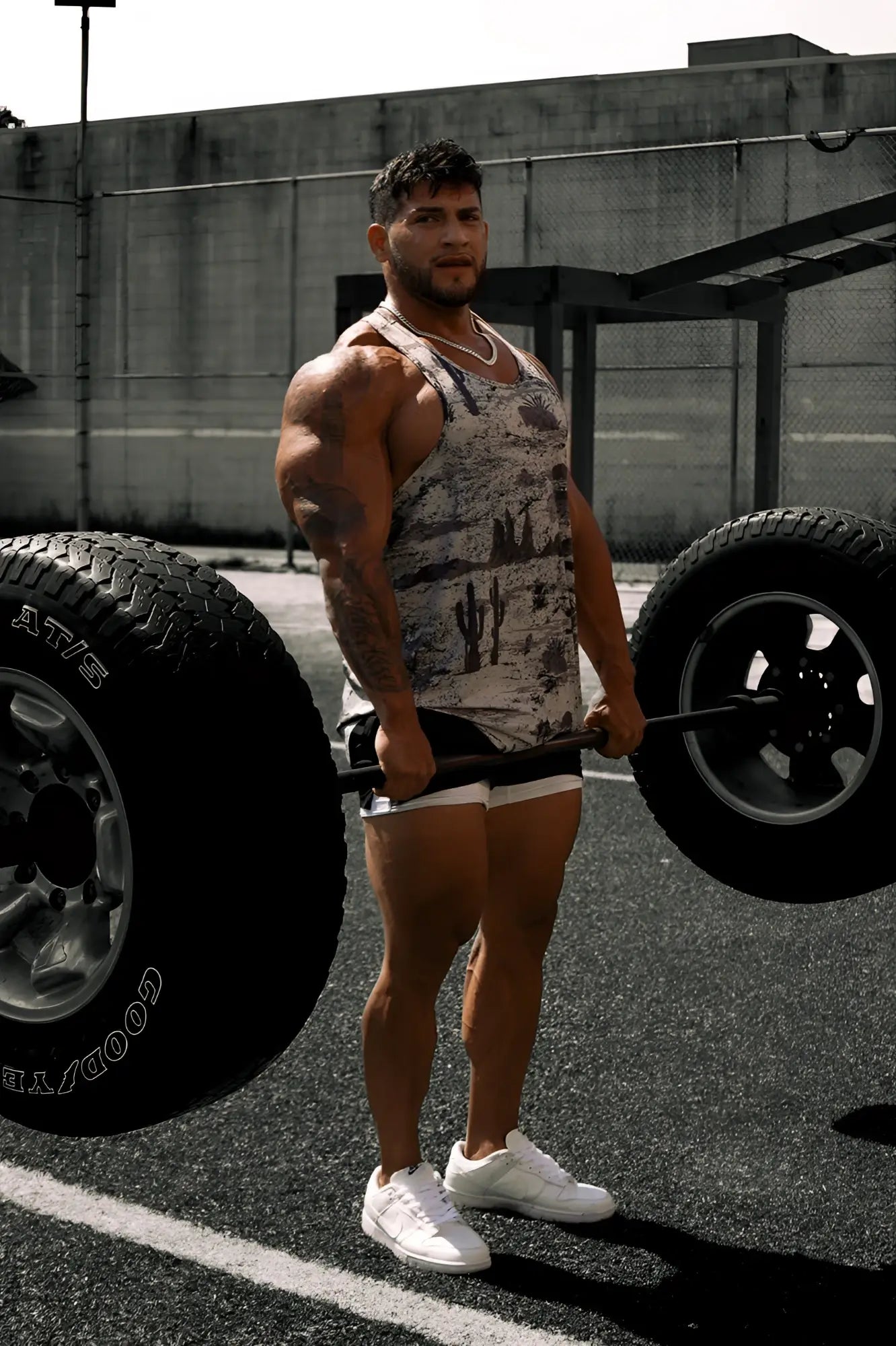 A muscular man wearing a gray and white cactus patterned graphic muscle stringer lifting a barbell with large tires