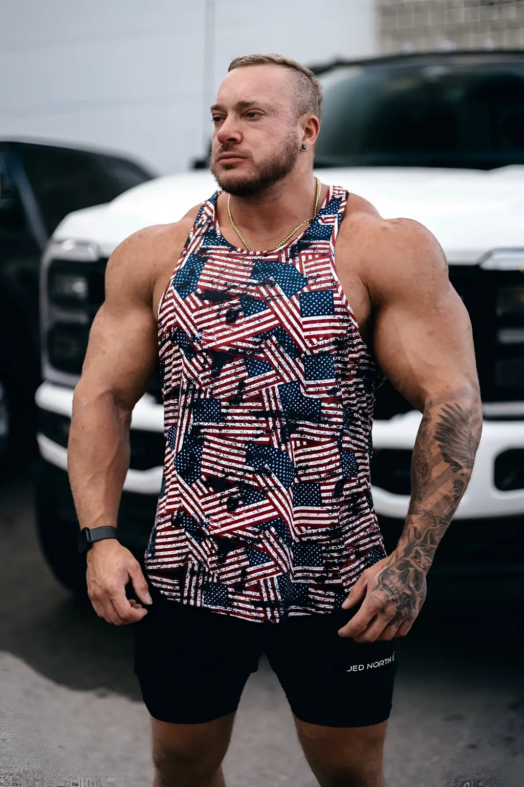 A muscular man wearing a fitted graphic muscle stringer tank top with a red white and blue USA flag pattern