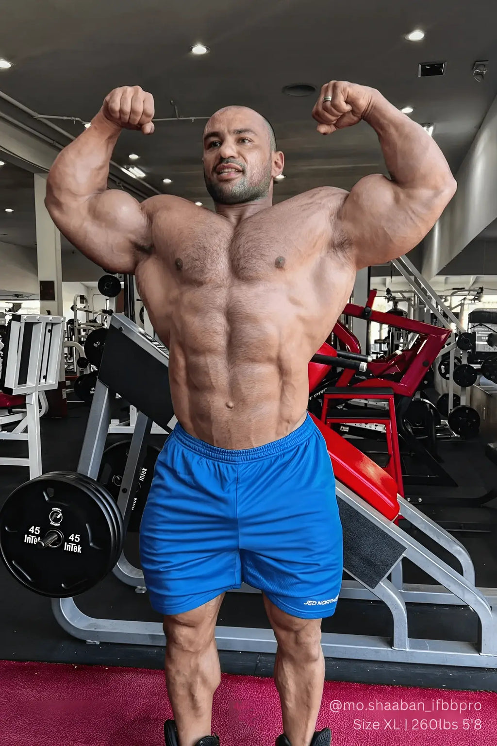 A muscular man flexing in a gym wearing blue Jed North Jersey Mesh 7.5 inch athletic gym shorts with jdi logo