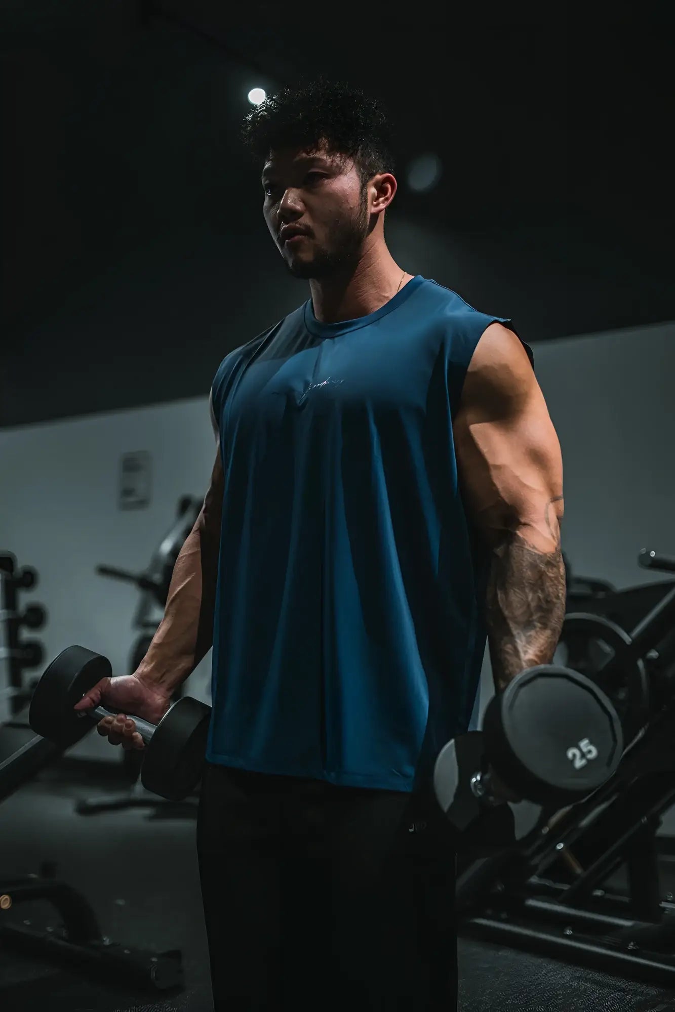 A man wearing a navy blue JN active muscle tee with a sleeveless design holding black dumbbells in a gym
