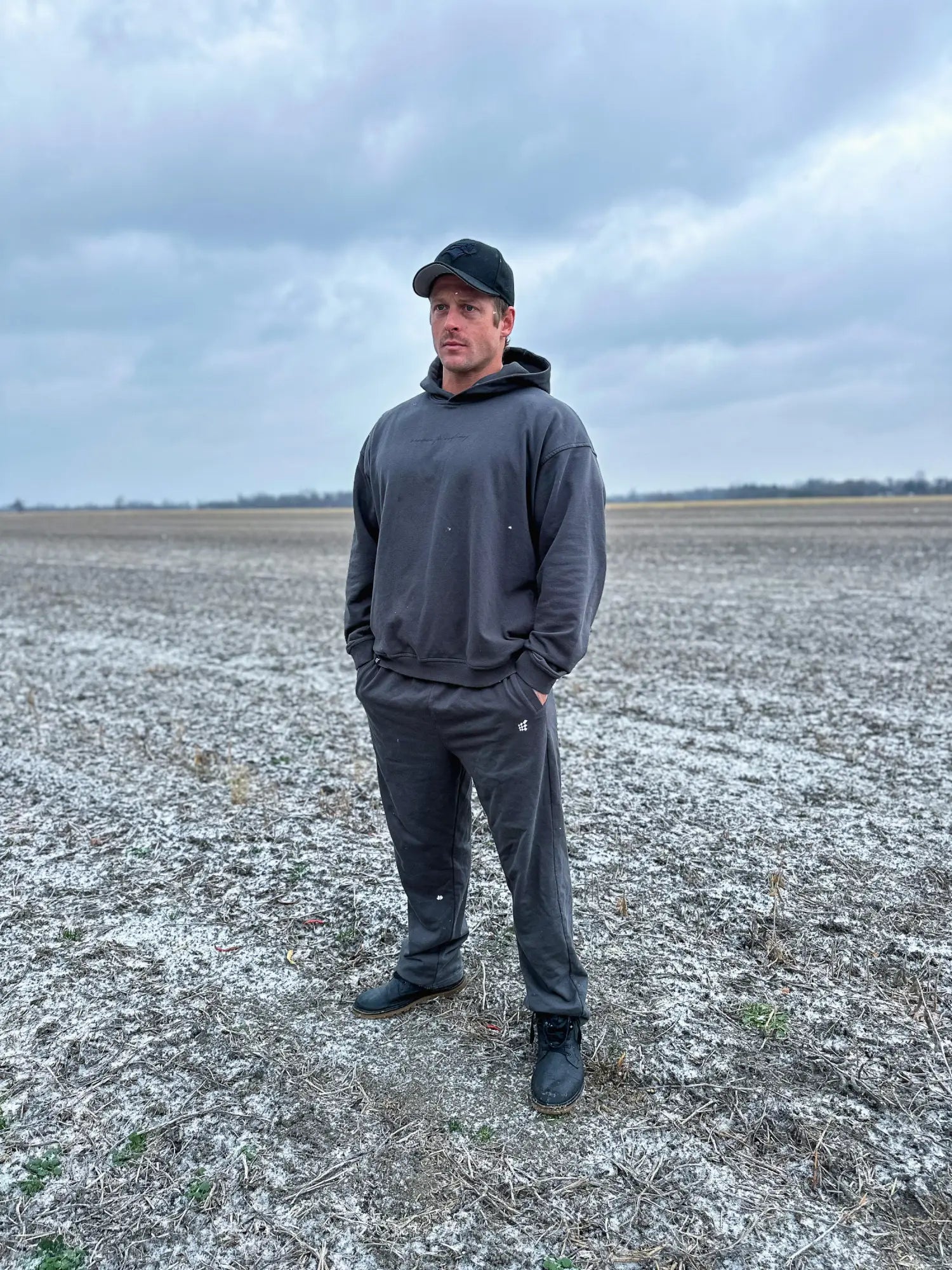 A man wearing gray jed north lax straight cut joggers and a dark gray hooded sweatshirt stands in an open field