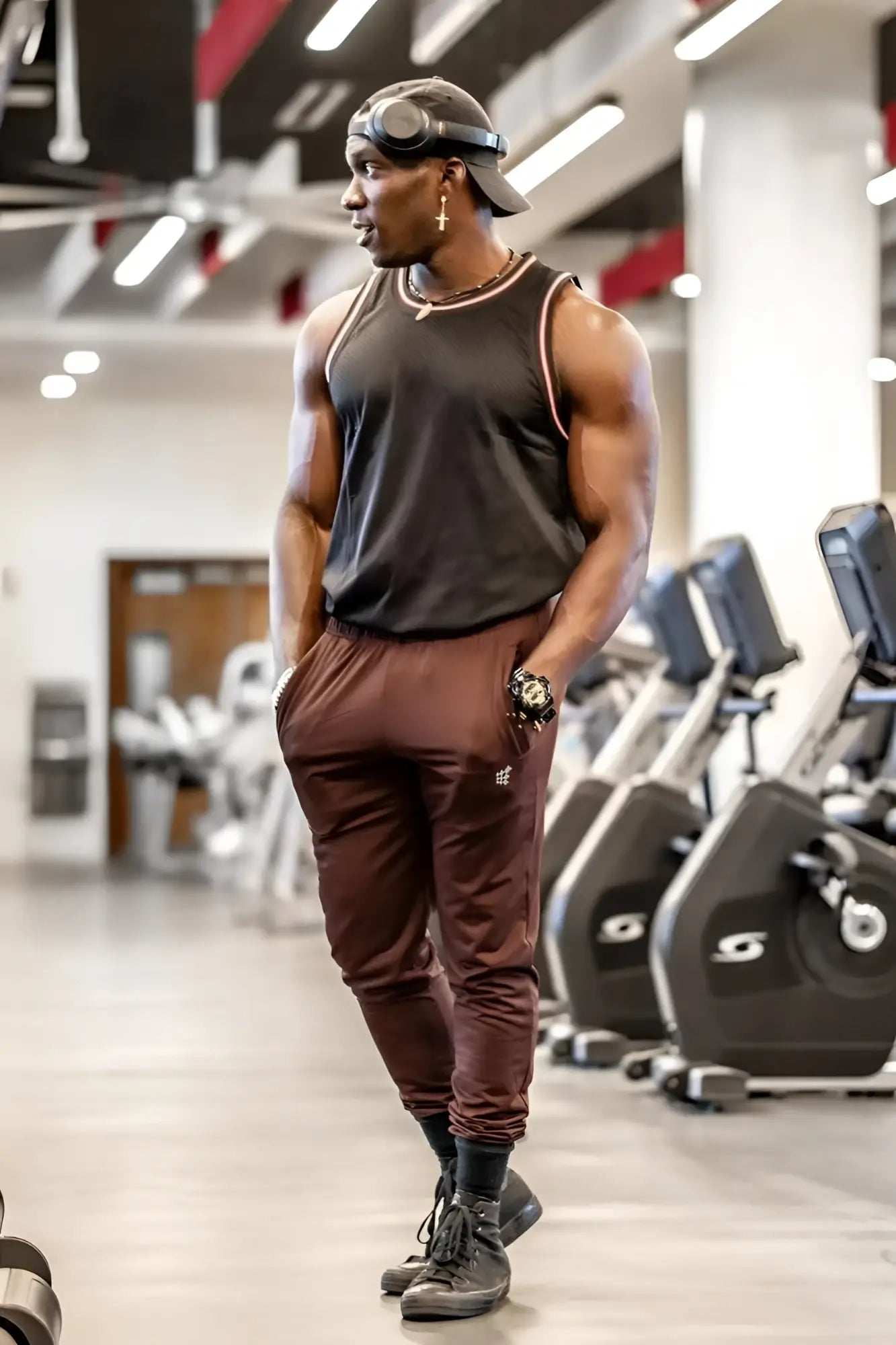 A muscular man wearing jed north matrix tapered brown joggers in maroon with a black sleeveless tank top and dark shoes