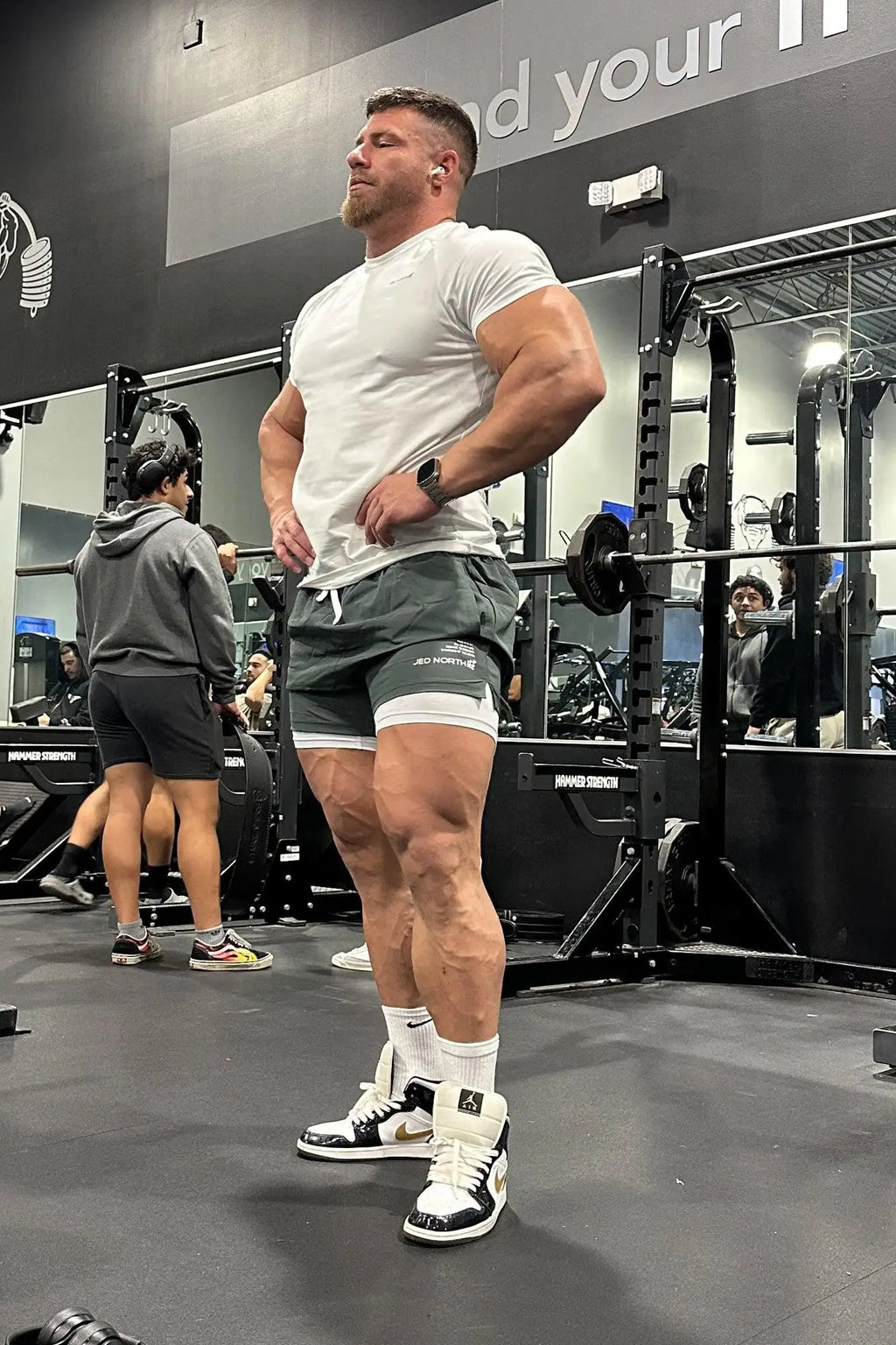 A muscular man wearing jed north mens 2-in-1 active 5 inch training shorts 2.0 in olive with a white t-shirt and black sneakers