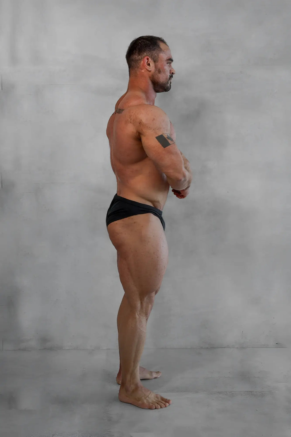 A muscular man stands in profile wearing black classic physique trunks 2 inch seam designed for men’s fitness
