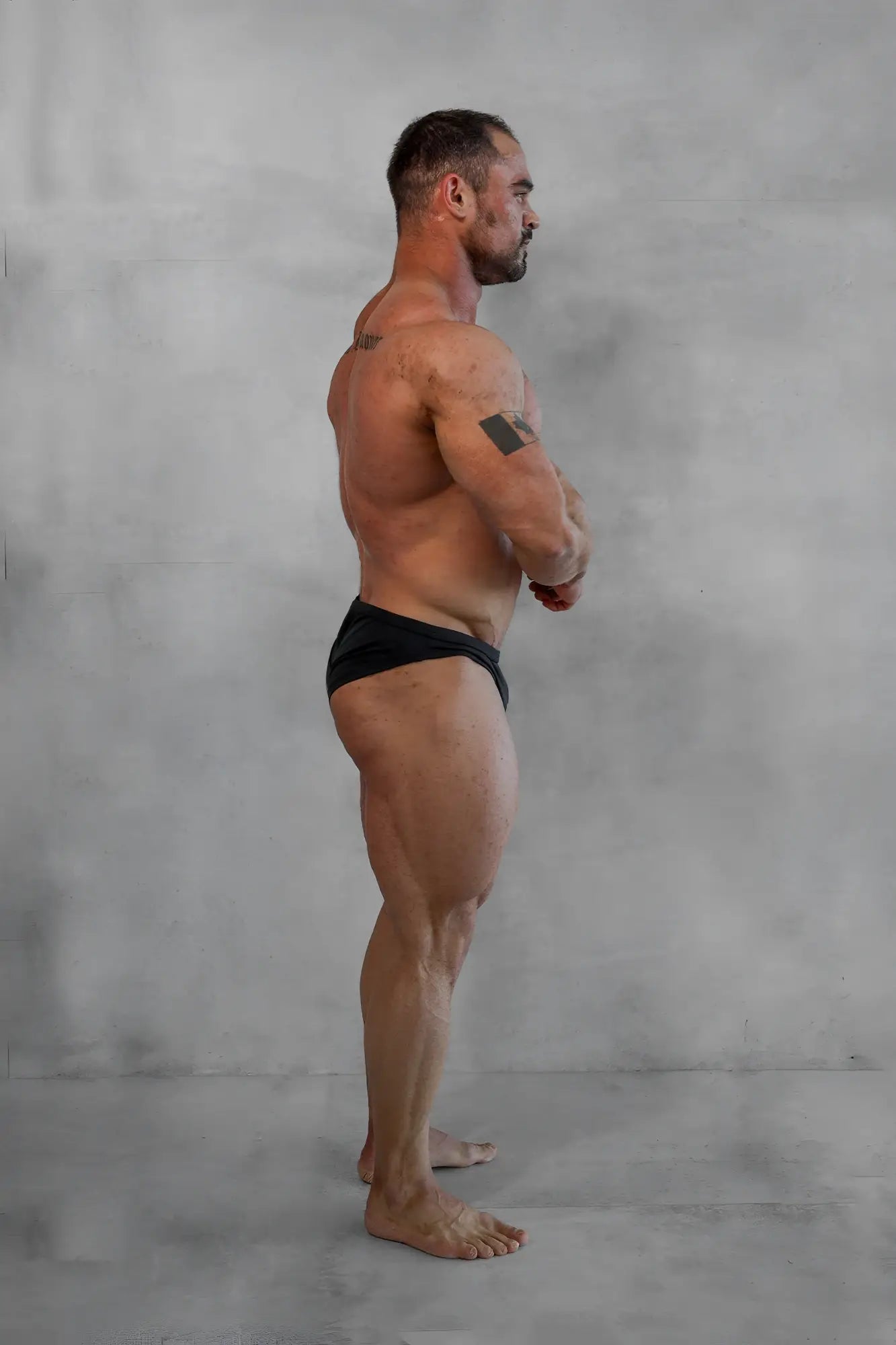 A muscular man stands in profile wearing black classic physique trunks 2 inch seam designed for men’s fitness