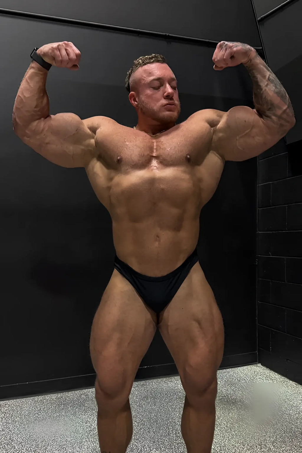 A muscular man with tattoos flexing his arms wearing black men’s classic physique trunks 2 inch seam briefs