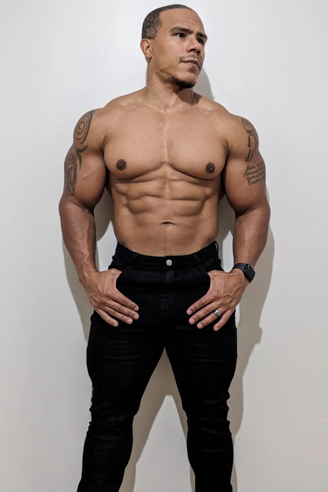 A muscular man wearing men’s fitted stretchy black jeans with a silver button and a black watch on his left wrist