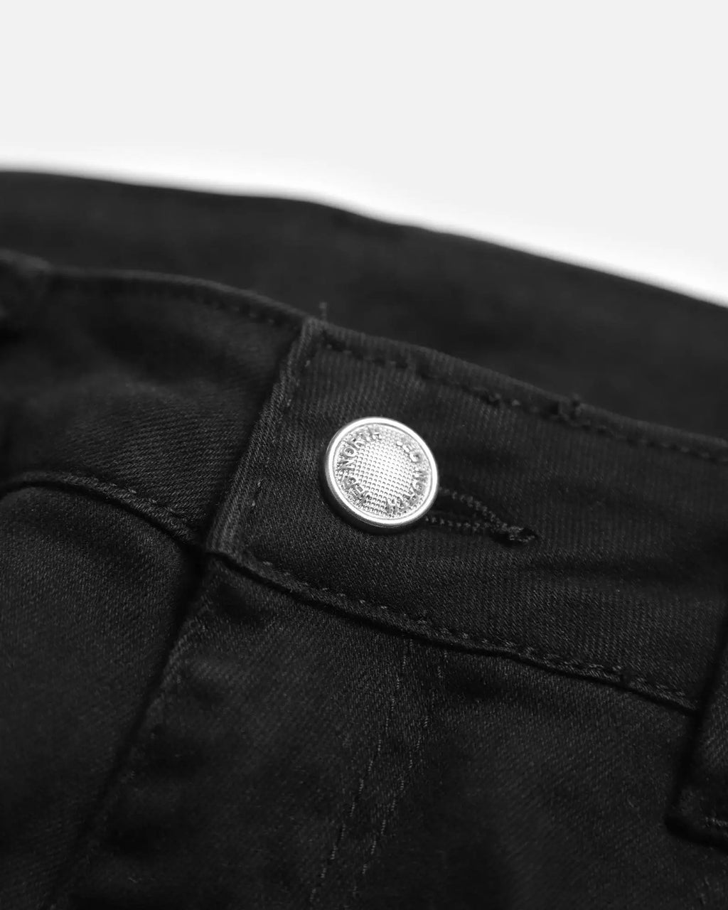 Silver button with engraved text on black men’s fitted stretchy pants made from dark denim fabric