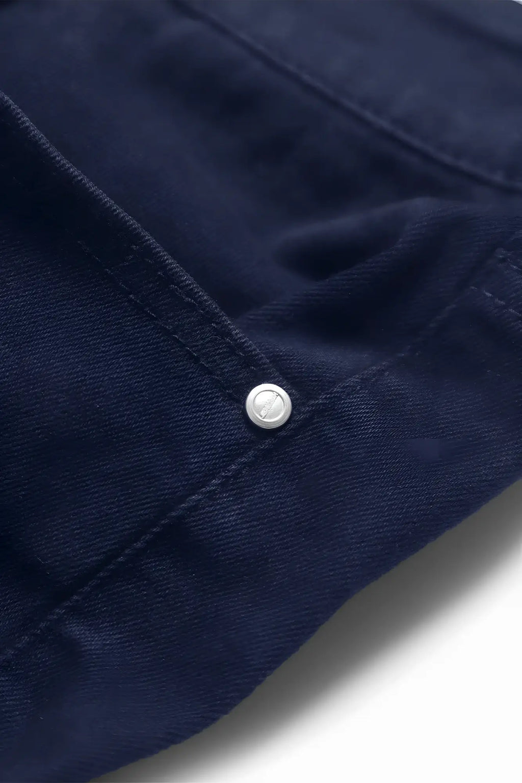A silver-toned button with embossed logo on navy stretchy denim fabric of men’s fitted stretchy pants back side