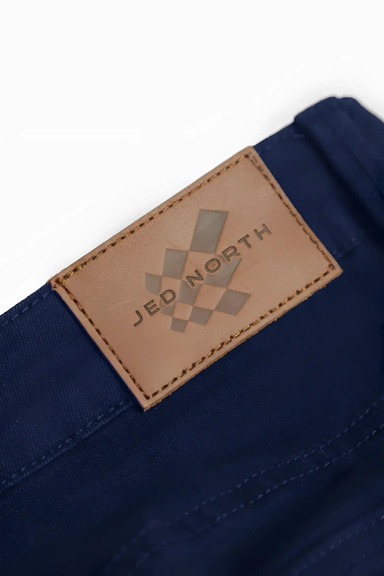 A tan leather patch with a debossed jed north logo stitched onto the back of men’s fitted stretchy navy denim pants