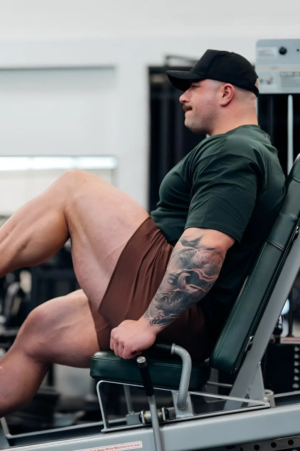 A man with a tattooed arm sits on a green and silver weight machine wearing jed north motion 5 inch varsity sweat shorts dark brown