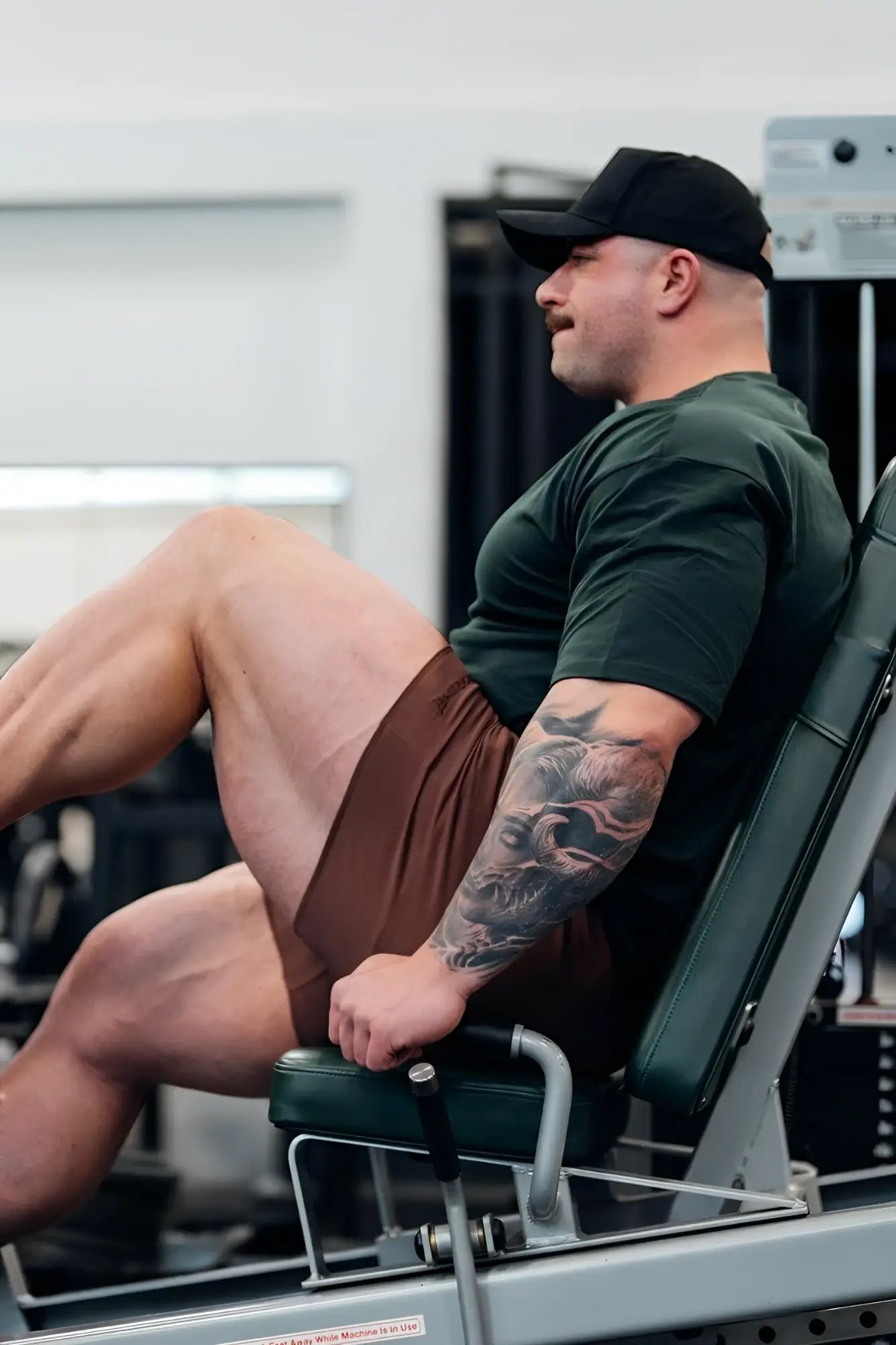 A man with a tattooed arm sits on a green and silver weight machine wearing jed north motion 5 inch varsity sweat shorts dark brown