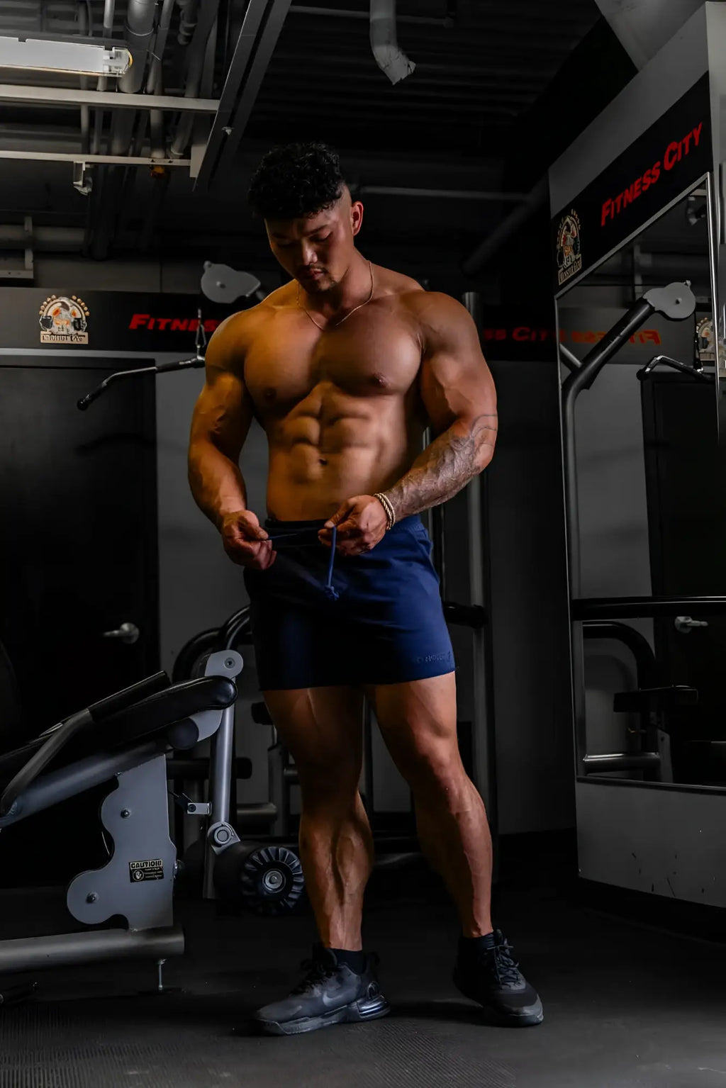 A muscular man wearing jed north motion 5 inch varsity sweat shorts in navy and black sneakers stands in a gym