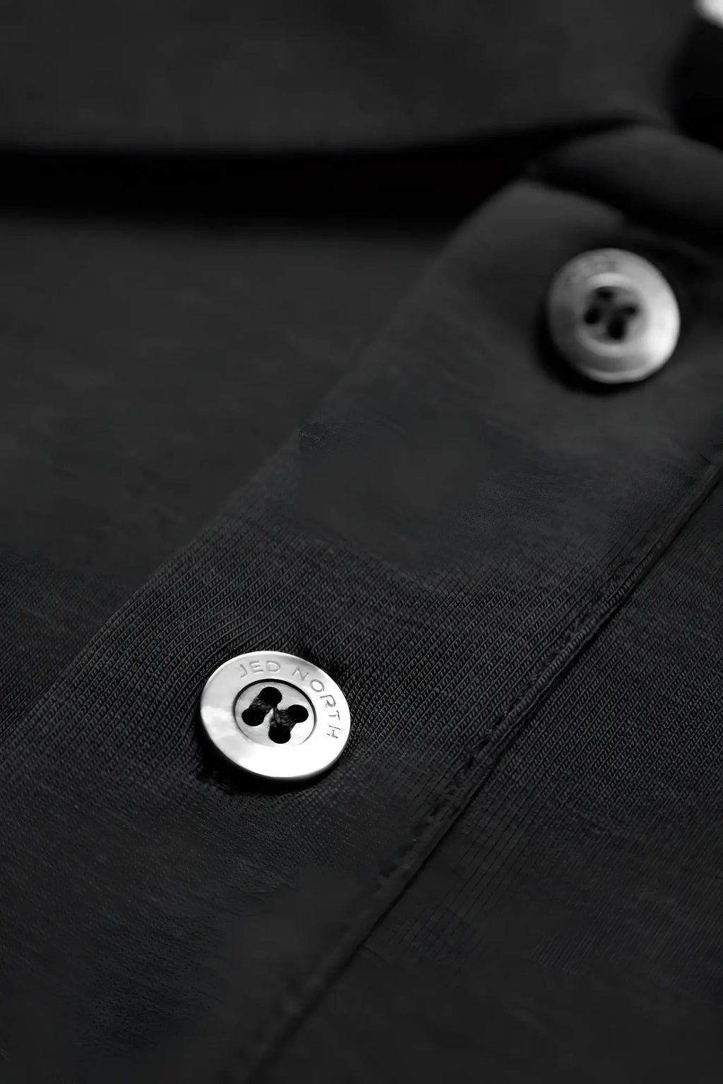 Silver button engraved with jed norris on the black fabric collar of jed north muscle fit all-day polo t-shirt onyx black