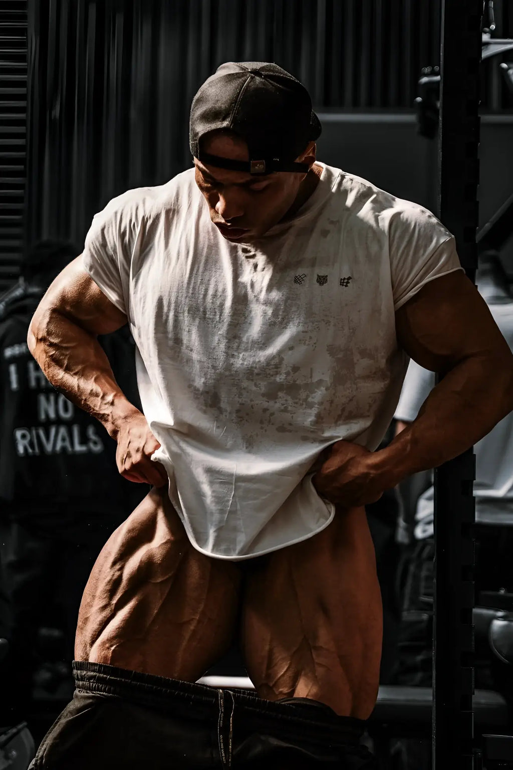 A muscular man wearing an oversized raw-cut cropped white tee with black logos on the chest and a dark backward cap