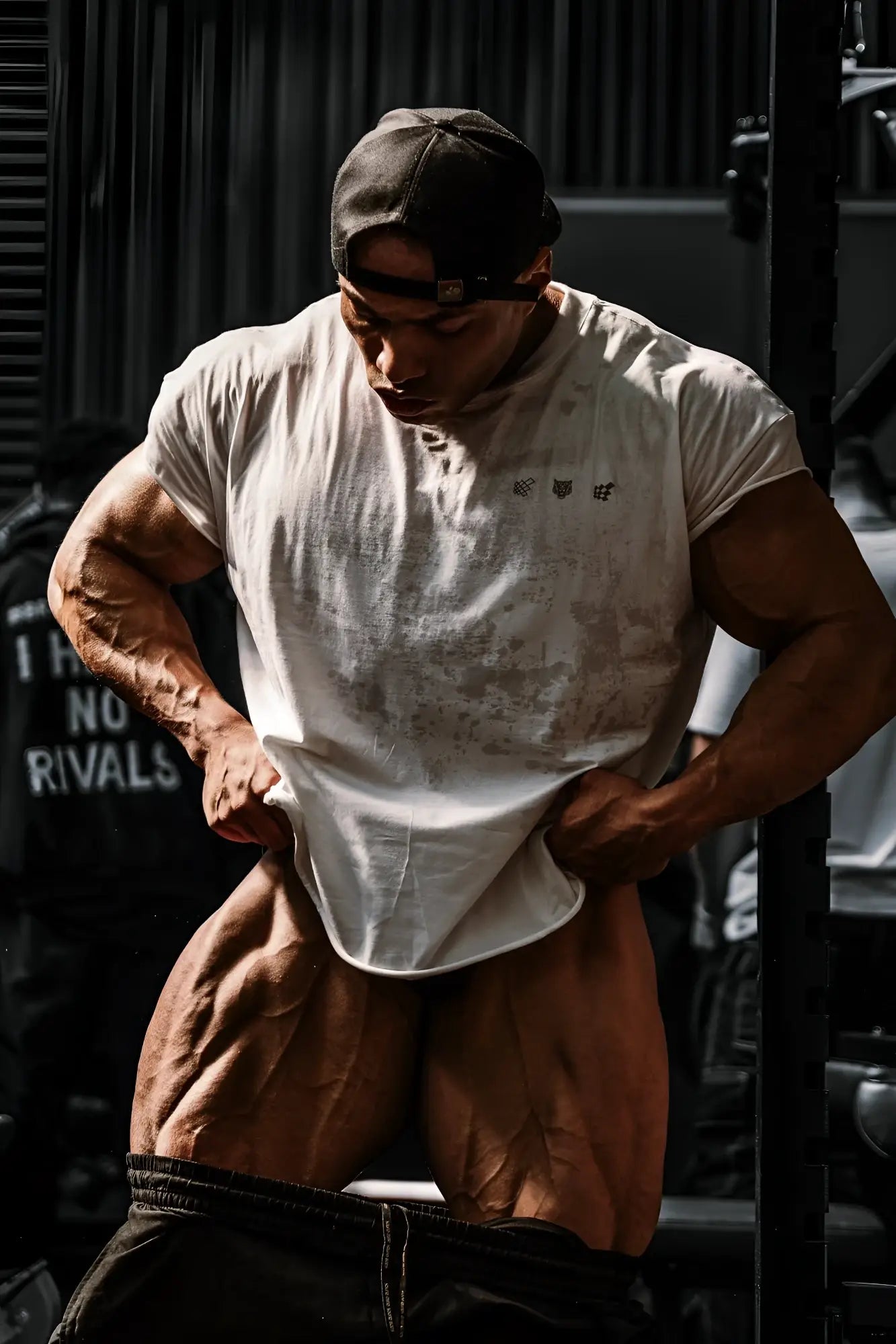 A muscular man wearing an oversized raw-cut cropped white tee with black logos on the chest and a dark backward cap