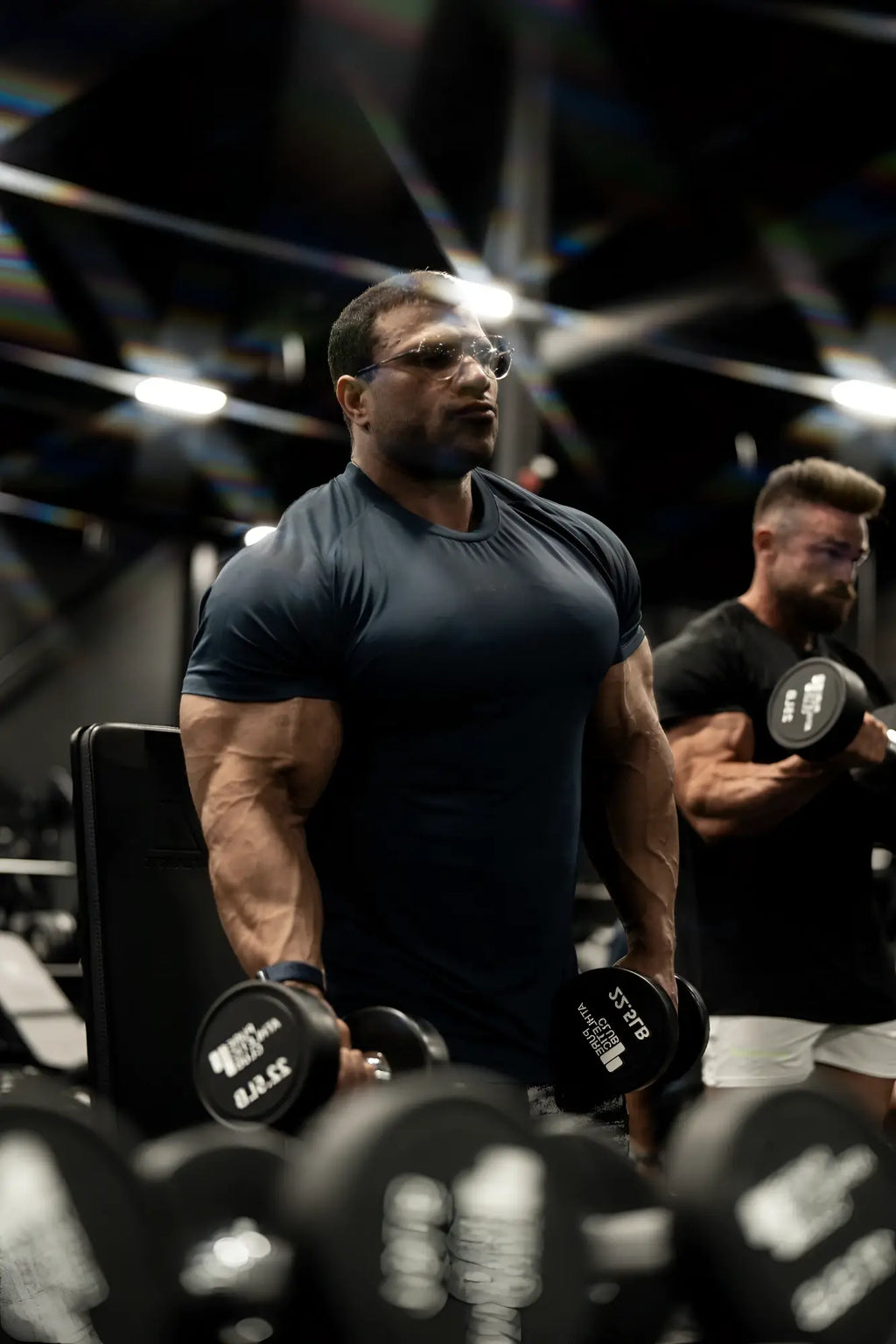 A muscular man wearing a black short-sleeved premium training tee holding dumbbells for strength training