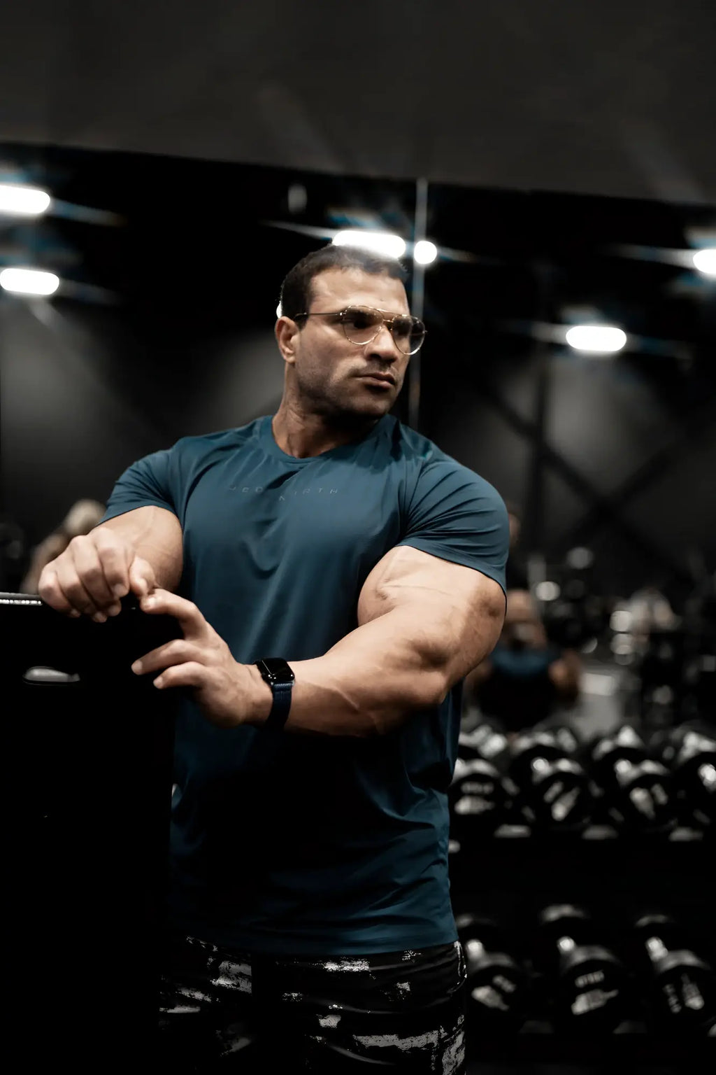 A muscular man wearing a navy premium training tee and patterned black pants stands in a gym with dumbbells behind him