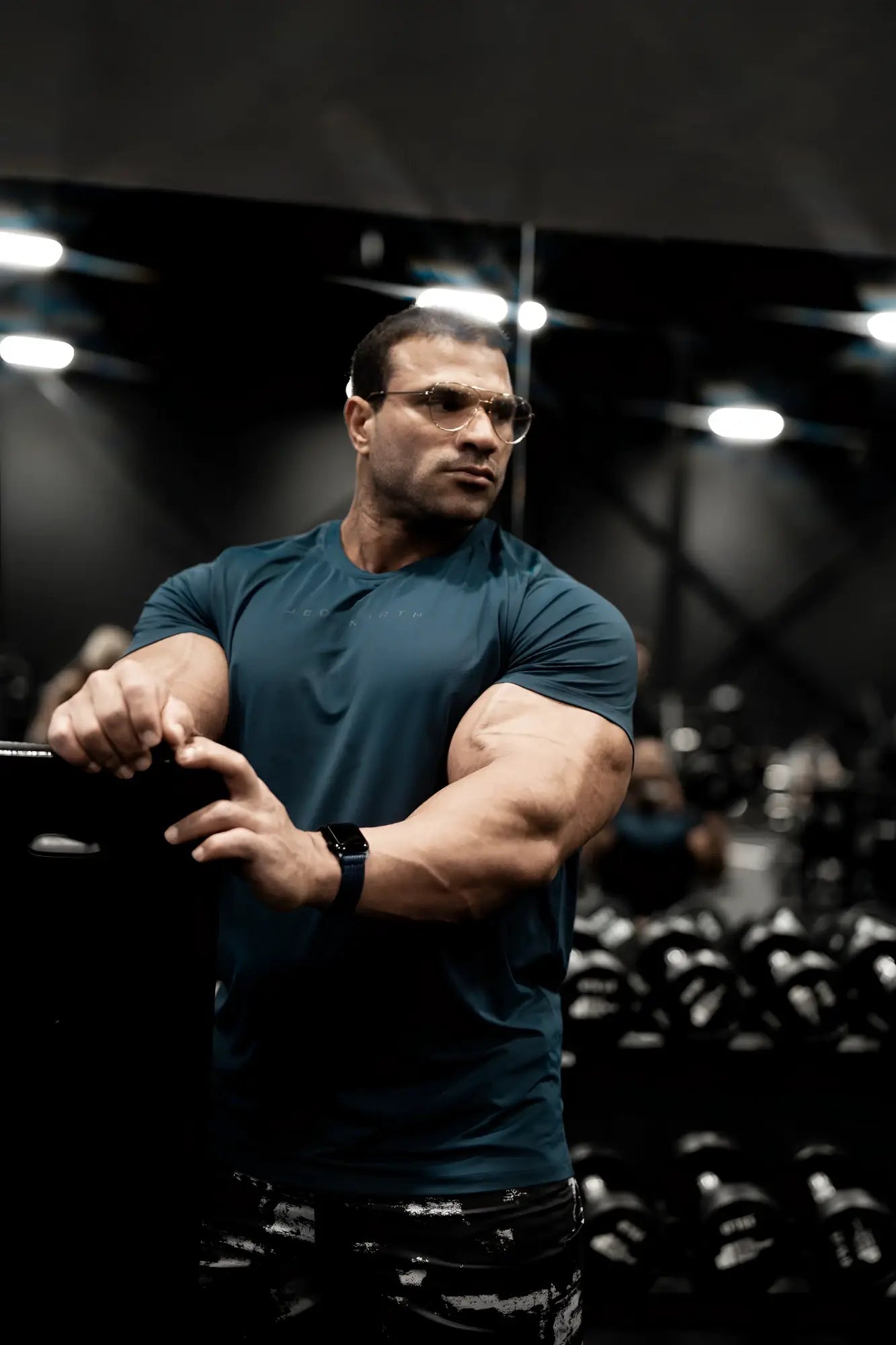 A muscular man wearing a navy premium training tee and patterned black pants stands in a gym with dumbbells behind him